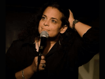 Comedian Jessica Kirson in a black shirt holding a microphone brushing hair off her face.