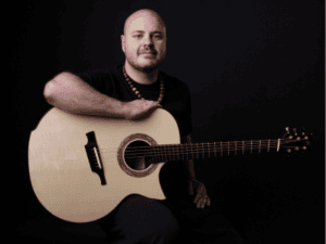 Andy McKee against black, holding his acoustic guitar 